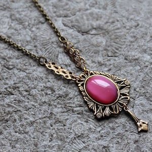 Könnte beinhalten: Eine goldene Halskette mit einem ovalen rosa Edelstein-Anhänger, der in einen verzierten Rahmen gefasst ist. Der Anhänger hat ein zartes, florales Design und hängt an einer Kette mit einem zarten, filigranen Muster.