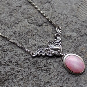 Könnte beinhalten: Eine silberne Halskette mit einem filigranen, verzierten Design zeigt einen ovalen rosa Edelstein-Anhänger. Die Halskette wird auf einer strukturierten grauen Oberfläche präsentiert.