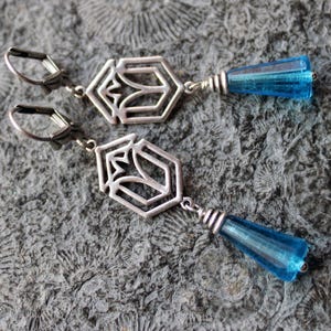 Könnte beinhalten: Ein Paar silberfarbene Ohrhänger mit geometrischem Design und blauen, tropfenförmigen Glasperlen. Die Ohrringe werden auf einer strukturierten, grauen Oberfläche präsentiert.