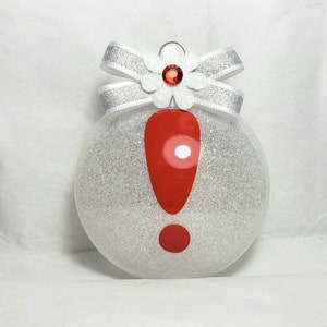 May include: A white glitter Christmas ornament with a red exclamation point and a white bow with a red jewel in the center.