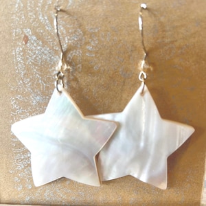 Op de afbeelding: Een paar stervormige oorbellen. De oorbellen hebben een parelmoerachtige, witte afwerking met een subtiele iriserende glans. De sterren zijn opgehangen aan goudkleurige oorhaken. De oorbellen zijn ontworpen om te bungelen en het licht te vangen.