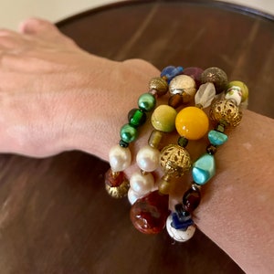 May include: A stack of colourful beaded bracelets with green, yellow, white, blue and brown beads. The bracelets are layered on a person's wrist.