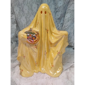 May include: A ceramic ghost figurine with a glossy yellow finish and a delicate floral pattern. The ghost features two hollow eye openings and holds a small, iridescent, metallic-toned pumpkin in its right arm, creating a decorative piece.