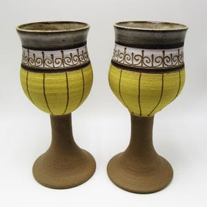 May include: Two ceramic wine goblets. The goblets have a yellow body with vertical brown lines, a decorative white and brown band, and a gray rim. The base and stem are brown.