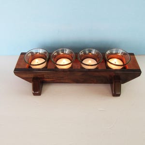 May include: A rectangular dark brown wooden candle holder featuring four circular cutouts, each containing a clear glass votive cup with a lit white tea light candle. The base is supported by two small wooden feet on either side.