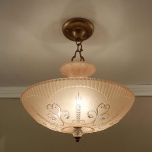 May include: A ceiling-mounted light fixture featuring a wide, pale pink, ribbed glass bowl shade. The shade is decorated with silver-toned scrollwork and floral accents. It is suspended by a brass-colored metal canopy and chain assembly.