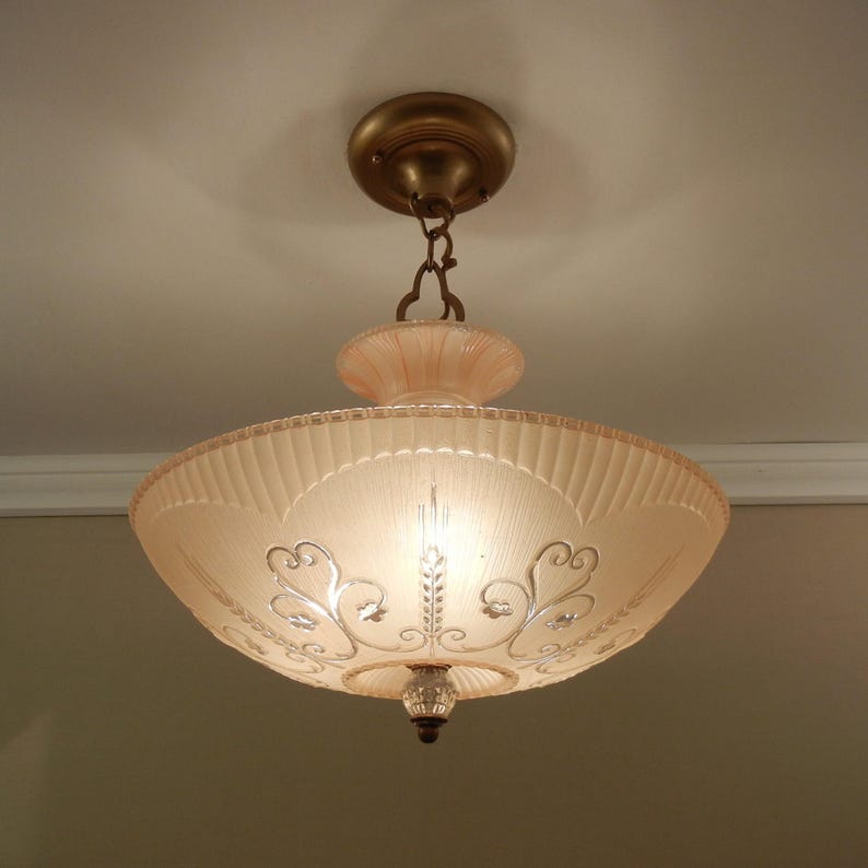 May include: A ceiling-mounted light fixture featuring a wide, pale pink, ribbed glass bowl shade. The shade is decorated with silver-toned scrollwork and floral accents. It is suspended by a brass-colored metal canopy and chain assembly.