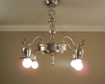Pode incluir: Um lustre de metal prateado polido com uma coluna central, uma base circular e quatro braços curvos. Cada braço suporta uma cúpula em forma de sino voltada para baixo com uma lâmpada branca brilhante e luminosa na parte inferior.