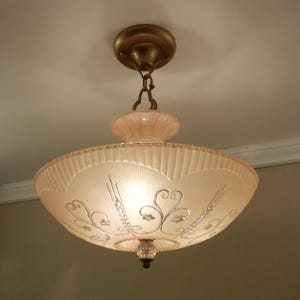 Pode incluir: Uma lumin&aacute;ria de teto com uma c&uacute;pula de vidro fosco em tom rosa p&aacute;lido, apresentando uma borda recortada e padr&otilde;es de volutas em relevo. A lumin&aacute;ria &eacute; suspensa por uma corrente e canopla de metal cor de bronze, com um pequeno remate de vidro.