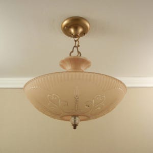 Pode incluir: Um candeeiro de teto semi-embutido com uma ta&ccedil;a de vidro fosco em tom de p&ecirc;ssego, decorada com volutas gravadas e estrias verticais. A pe&ccedil;a inclui uma base de metal dourado, uma corrente curta e um pequeno remate de vidro transparente.