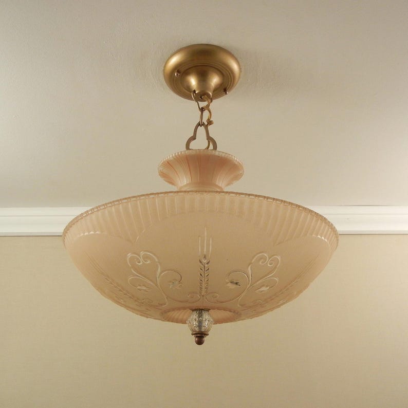 Pode incluir: Um candeeiro de teto semi-embutido com uma ta&ccedil;a de vidro fosco em tom de p&ecirc;ssego, decorada com volutas gravadas e estrias verticais. A pe&ccedil;a inclui uma base de metal dourado, uma corrente curta e um pequeno remate de vidro transparente.