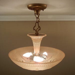 Pode incluir: Uma lumin&aacute;ria de teto com c&uacute;pula de vidro canelado em rosa p&aacute;lido e delicados padr&otilde;es florais gravados. A lumin&aacute;ria &eacute; suspensa por uma corrente e canopla de metal em tom bronze, contendo tr&ecirc;s l&acirc;mpadas acesas em seu interior.