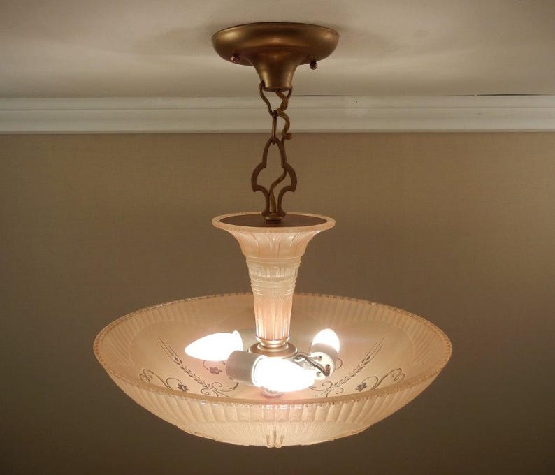 Pode incluir: Uma lumin&aacute;ria de teto com c&uacute;pula de vidro canelado em rosa p&aacute;lido e delicados padr&otilde;es florais gravados. A lumin&aacute;ria &eacute; suspensa por uma corrente e canopla de metal em tom bronze, contendo tr&ecirc;s l&acirc;mpadas acesas em seu interior.