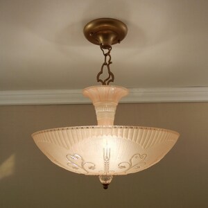 Pode incluir: Uma lumin&aacute;ria de teto com uma c&uacute;pula de vidro fosco em rosa p&aacute;lido, apresentando um design canelado e padr&otilde;es de volutas gravados. A lumin&aacute;ria &eacute; suspensa por uma corrente e canopla de metal cor de bronze, com um pequeno remate de vidro claro.