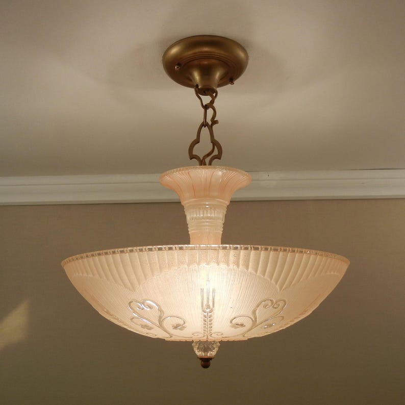 Pode incluir: Uma lumin&aacute;ria de teto com uma c&uacute;pula de vidro fosco em rosa p&aacute;lido, apresentando um design canelado e padr&otilde;es de volutas gravados. A lumin&aacute;ria &eacute; suspensa por uma corrente e canopla de metal cor de bronze, com um pequeno remate de vidro claro.