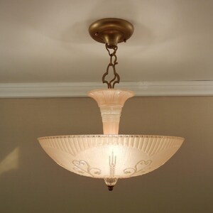 Pode incluir: Uma lumin&aacute;ria de teto com uma ta&ccedil;a de vidro fosco rosa p&aacute;lido com padr&otilde;es gravados de leques e volutas. A ta&ccedil;a &eacute; suspensa por uma corrente de metal cor de bronze e uma canopla circular correspondente, com um suporte central de vidro.