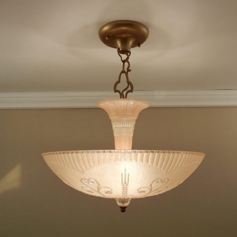 Pode incluir: Uma lumin&aacute;ria de teto com uma ta&ccedil;a de vidro fosco rosa p&aacute;lido com padr&otilde;es gravados de leques e volutas. A ta&ccedil;a &eacute; suspensa por uma corrente de metal cor de bronze e uma canopla circular correspondente, com um suporte central de vidro.