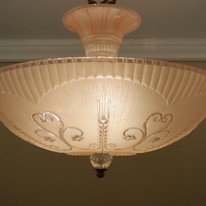 Pode incluir: Uma lumin&aacute;ria de teto de vidro rosa p&aacute;lido com formato de tigela larga e rasa, borda canelada e padr&otilde;es florais em relevo transparentes. A haste central tamb&eacute;m &eacute; feita de vidro rosa correspondente, e um pequeno remate de vidro transparente pende do centro inferior da tigela.