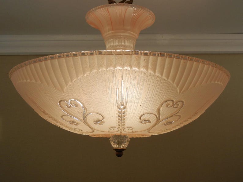 Pode incluir: Uma lumin&aacute;ria de teto de vidro rosa p&aacute;lido com formato de tigela larga e rasa, borda canelada e padr&otilde;es florais em relevo transparentes. A haste central tamb&eacute;m &eacute; feita de vidro rosa correspondente, e um pequeno remate de vidro transparente pende do centro inferior da tigela.