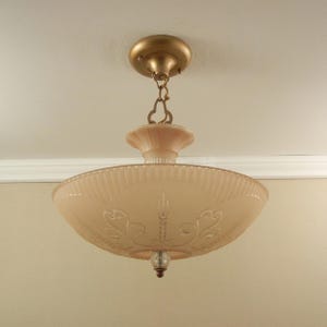 Pode incluir: Uma lumin&aacute;ria de teto com uma c&uacute;pula de vidro fosco em rosa p&aacute;lido, apresentando padr&otilde;es gravados de volutas e espigas. A pe&ccedil;a inclui uma base de metal em tom de lat&atilde;o, uma corrente decorativa e um pequeno remate de vidro transparente.