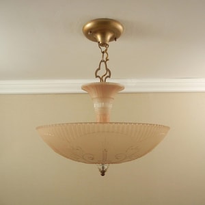Pode incluir: Uma lumin&aacute;ria de teto com uma c&uacute;pula de vidro canelado em rosa p&aacute;lido e detalhes florais gravados. A lumin&aacute;ria &eacute; suspensa por uma corrente e canopla de metal dourado, com uma haste central de vidro e um pequeno remate decorativo na base.