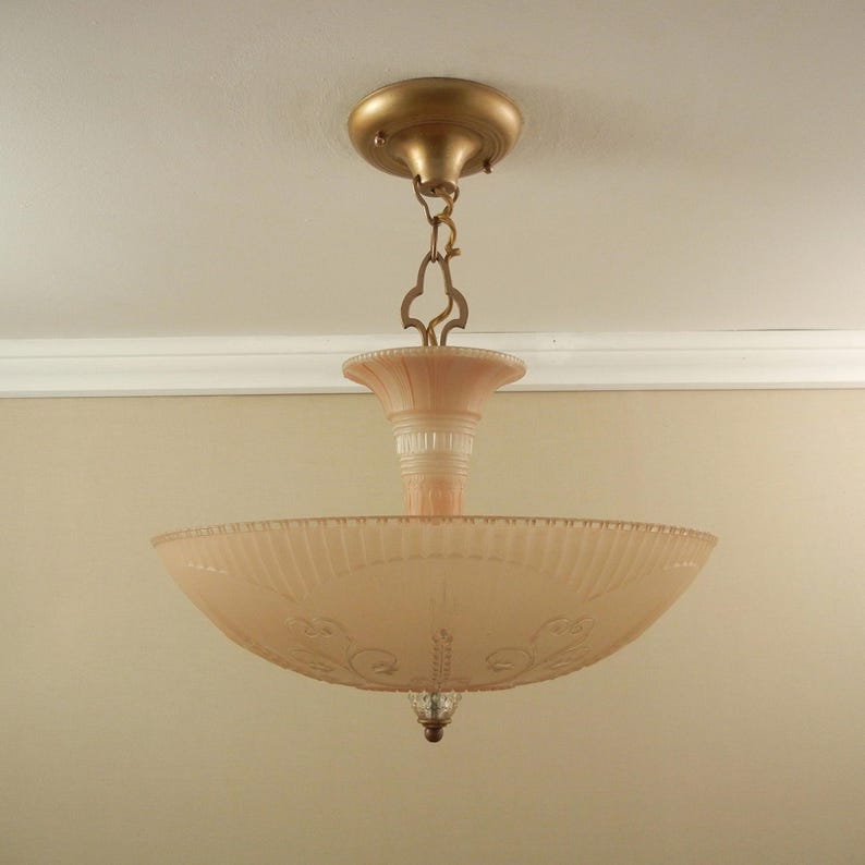 Pode incluir: Uma lumin&aacute;ria de teto com uma c&uacute;pula de vidro canelado em rosa p&aacute;lido e detalhes florais gravados. A lumin&aacute;ria &eacute; suspensa por uma corrente e canopla de metal dourado, com uma haste central de vidro e um pequeno remate decorativo na base.