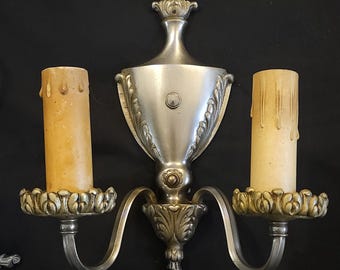 Pode incluir: Arandela de parede prateada com dois castiçais. A arandela apresenta detalhes ornamentados, incluindo detalhes florais e um topo decorativo. Os castiçais curvos seguram velas creme, adicionando um brilho quente ao design.