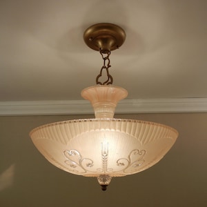 Pode incluir: Uma lumin&aacute;ria de teto suspensa com uma c&uacute;pula de vidro fosco em rosa p&aacute;lido, borda canelada e padr&otilde;es de volutas gravados. A luz &eacute; suspensa por uma corrente e canopla de metal cor de lat&atilde;o, finalizada com um pequeno remate de vidro.