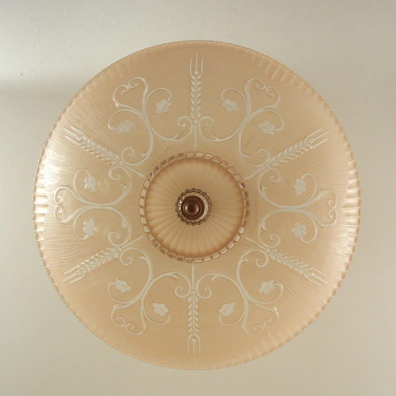Pode incluir: Uma lumin&aacute;ria de teto redonda de vidro cor de p&ecirc;ssego com uma c&uacute;pula central canelada e um remate castanho escuro. O abajur de vidro &eacute; decorado com espigas de trigo e padr&otilde;es de volutas gravados em branco, com uma borda exterior estriada.