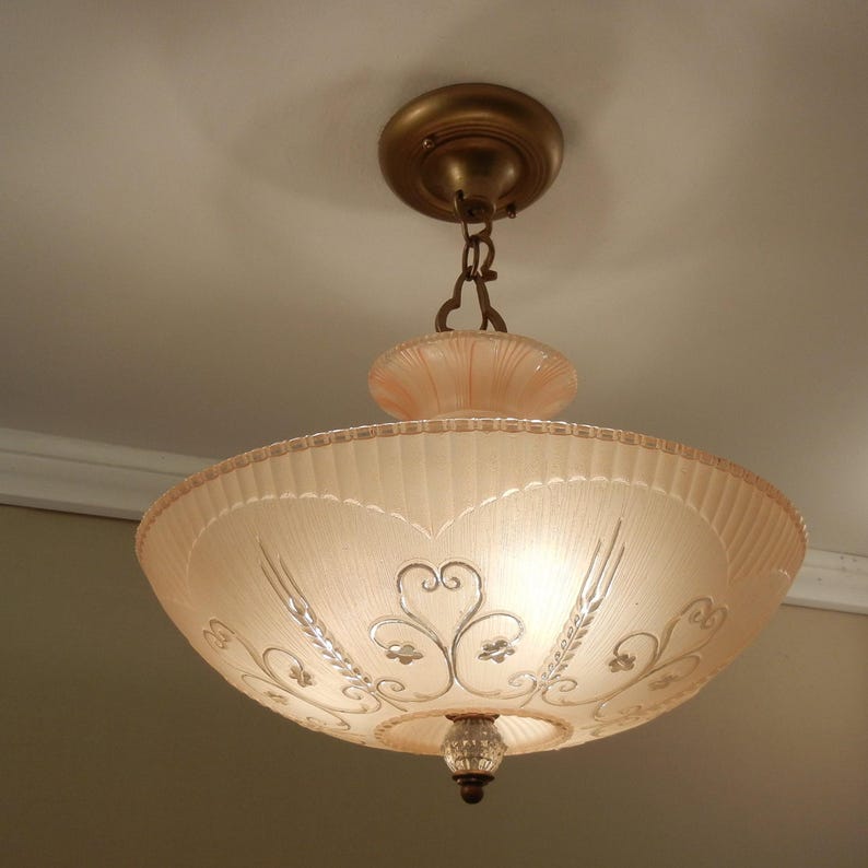 Pode incluir: Uma lumin&aacute;ria de teto com uma c&uacute;pula de vidro em forma de ta&ccedil;a, de cor rosa p&aacute;lido, com textura canelada e padr&otilde;es de volutas prateadas. A lumin&aacute;ria est&aacute; suspensa por uma corrente met&aacute;lica de uma base circular de cor bronze a condizer.