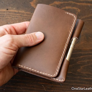 Op de afbeelding: Een bruine lederen notitieboekhoes met een gladde afwerking en dikke witte stiksels langs de randen. Een metalen pen is in een speciale lus aan de zijkant gestoken. De tekst OneStarLeatherGoods.com staat in de rechterbenedenhoek.