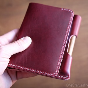Op de afbeelding: Een bordeauxrode leren notitieboekhoes met witte stiksels. De hoes heeft een penhouder en wordt in de hand van een persoon gehouden. De notitieboekhoes is gemaakt van hoogwaardig leer.