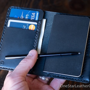 Op de afbeelding: Zwart leren portemonnee met kaartsleuven, een notitieblok en een pen. De portemonnee bevat een Visa en een Mastercard. Het notitieblok is zwart en de pen is zwart met een zilveren punt. De portemonnee is opengehouden.