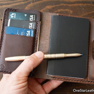 Op de afbeelding: Een donkerbruine lederen portemonnee met een notitieboekje en een goudkleurige metalen pen. De portemonnee heeft drie kaartsleuven met creditcards en een vak voor een klein zwart notitieboekje. Een ster-logo is linksonder in het leer gedrukt.