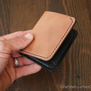 Op de afbeelding: Een bruine leren portemonnee met zwarte stiksels en een zwarte leren voering. De portemonnee is dubbelgevouwen en wordt in een hand gehouden.