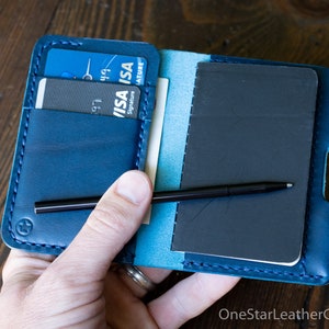 May include: Open blue leather wallet with a notebook, pen, and credit cards. The wallet is open, revealing a small black notebook and a black pen. Credit cards are visible in the card slots. The wallet is held open.