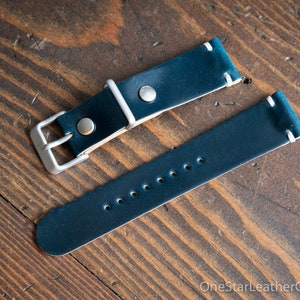 May include: A teal blue leather watch strap with white stitching and a silver buckle. The strap is laid out flat on a wooden surface.