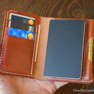 Op de afbeelding: Open bruin leren portemonnee met kaartsleuven en een notitieboekje. De portemonnee heeft witte stiksels en een messing scharnier. Creditcards zijn zichtbaar in de kaartsleuven. De tekst "OneStarLeatherGoods.com" staat rechtsonder.