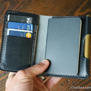 Op de afbeelding: Zwart leren portemonnee met kaartsleuven en een notitieboekje. De portemonnee is open en toont creditcards en een grijs notitieboekje. Aan de zijkant is een penhouder zichtbaar. De portemonnee wordt in een hand gehouden.