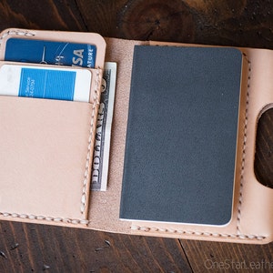 May include: A light tan leather wallet, open to reveal card slots with credit cards, a stack of dollar bills, and a small gray notebook. A gold-colored pen rests beside the wallet. The wallet is on a dark wood surface.