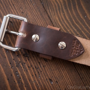 May include: A brown leather belt with a silver buckle and a brown leather strap. The strap has the text "ONE STAR 2016" embossed on it.