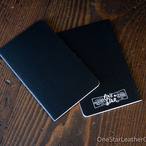 Könnte beinhalten: Zwei schwarze Notizbücher mit einem weißen Logo, das auf der Vorderseite "One Star Leather Goods" lautet.