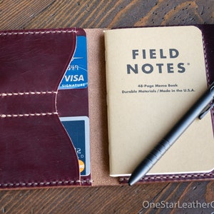 May include: Burgundy leather wallet with card slots, a Field Notes memo book, and a black pen. The wallet is open, displaying the contents. The memo book reads "FIELD NOTES" and "Made in the U.S.A."