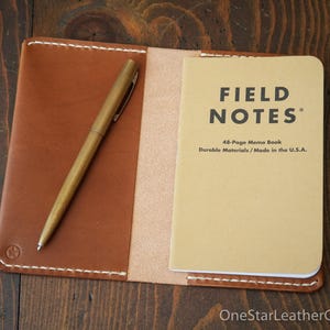 May include: Open leather notebook cover in brown with a brass pen and a Field Notes memo book. The memo book is tan with black text that reads "FIELD NOTES" and "48-Page Memo Book Durable Materials / Made in the U.S.A."