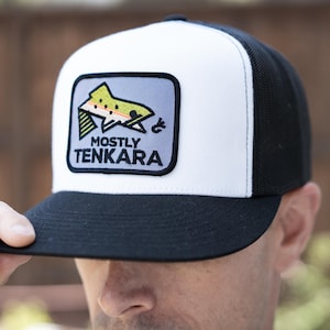 May include: A black and white trucker hat with a square patch featuring a stylized fish and the text "MOSTLY TENKARA". The hat has a white front panel, a black mesh back, and a black brim. The patch has a light blue background.