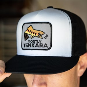 Puede incluir: Gorra de camionero en blanco y negro con un panel frontal blanco y una parte trasera de malla negra. La gorra presenta un parche rectangular con un gráfico de pez y el texto "MOSTLY TENKARA".