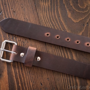May include: A brown leather belt with a silver buckle and five holes. The belt is shown on a wooden surface.
