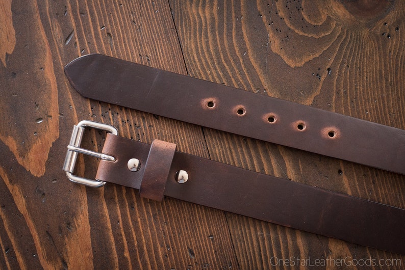 May include: A brown leather belt with a silver buckle and five holes. The belt is shown on a wooden surface.