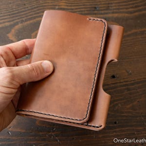 Puede incluir: Una cartera de cuero color canela con un acabado mate suave y costuras en contraste de color marrón oscuro a lo largo de los bordes. La cartera tiene forma rectangular con una sección recortada en el lado derecho y un diseño de solapa.