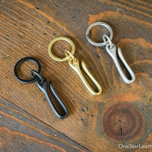 May include: Three keychains in black, gold, and silver tones rest on a wooden surface. Each keychain features a circular ring and a curved hook design. The wood grain is visible, adding a rustic touch.